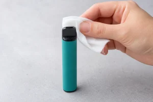 An adult hand wiping condensation from the outside of a disposable vape mouthpiece with a dry tissue